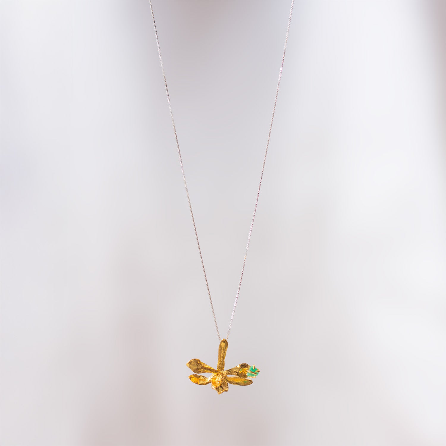 Collar con dije dorado de orquídea Prosthechea Sceptra real y pequeña esmeralda verde, suspendido de una fina cadena de plata sobre fondo claro.