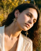 Modelo usando aretes largos de ramita con orquídea Oncidioides real inmortalizada en oro 24k y esmeraldas colombianas — joyería botánica Colombia, fundición orgánica, Liz de la Cruz
