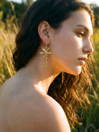 Aretes botánicos largos dorados con orquídea silvestre colombiana y esmeraldas naturales sobre ramita inmortalizada, usados en exterior, joyería botánica Bogotá, Liz de la Cruz
