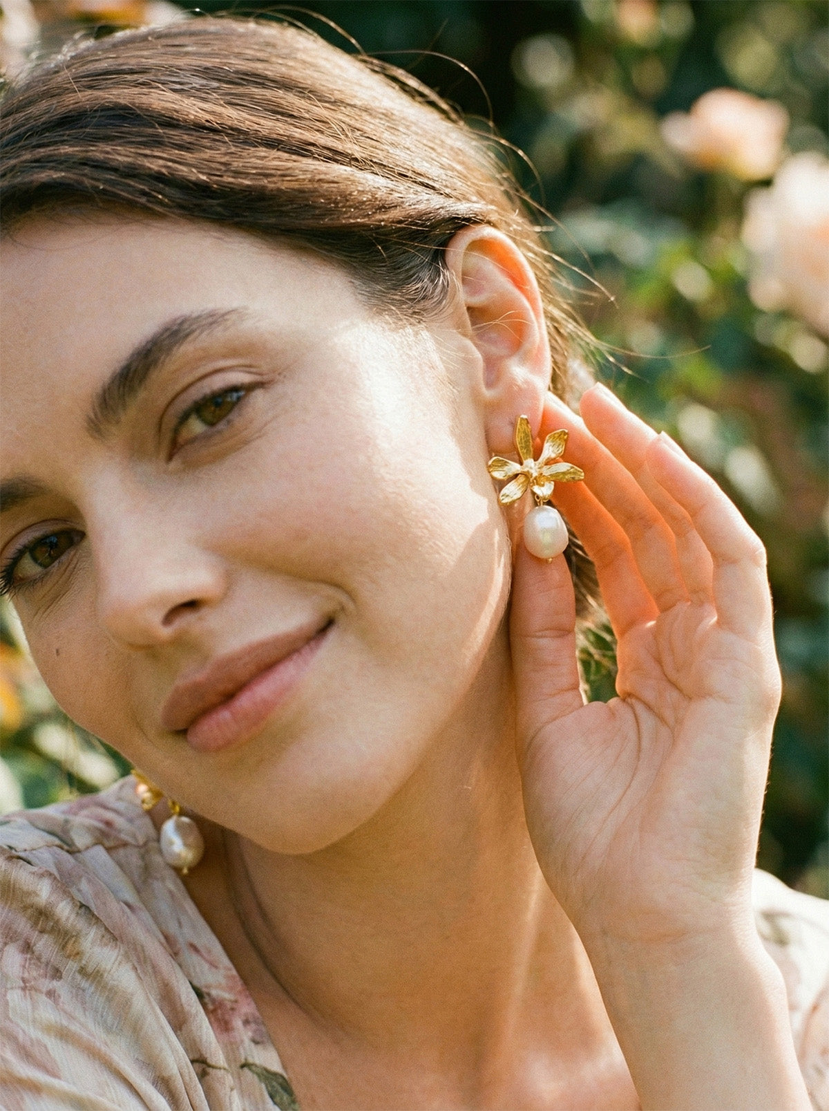 Modelo luciendo aretes de orquídea dorada y perla, resaltando el brillo orgánico y la elegancia del diseño artesanal en un entorno natural.