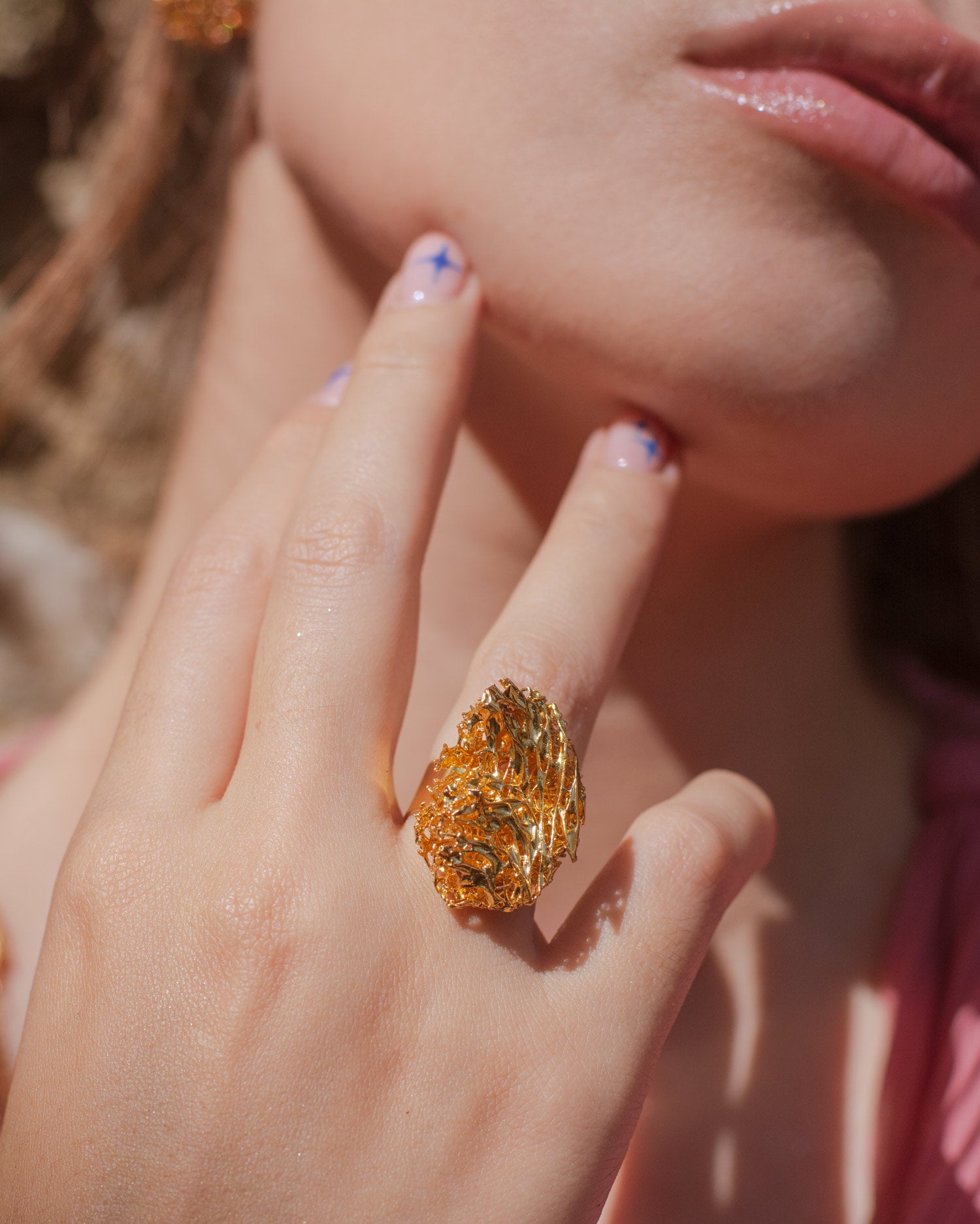 Anillo Luffa Estropajo Redondo de oro brillante en el dedo anular de una mano femenina, destacando su textura única y detallada inspirada en una esponja vegetal, bajo la luz del sol que realza su acabado metálico